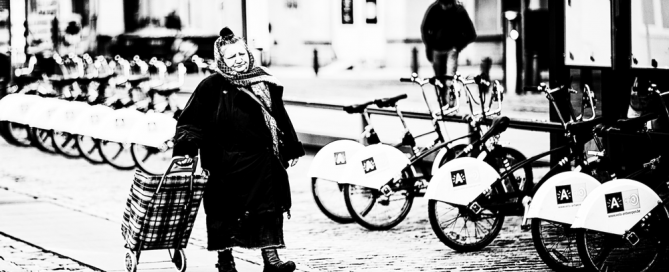 Eine ältere Frau mit Einkaufstrolley und Kopftuch geht an modernen Mieträdern vorbei