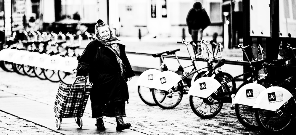 Eine ältere Frau mit Einkaufstrolley und Kopftuch geht an modernen Mieträdern vorbei