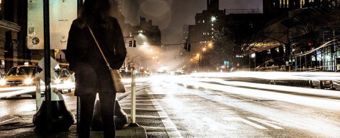 Eine Frau steht auf einer Verkehrsinsel inmitten einer vierspurigen Straße. Es ist dunkel und man sieht mehrere Autoscheinwerfer.