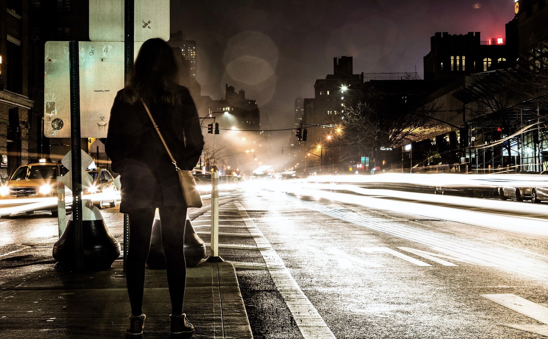 Eine Frau steht auf einer Verkehrsinsel inmitten einer vierspurigen Straße. Es ist dunkel und man sieht mehrere Autoscheinwerfer.